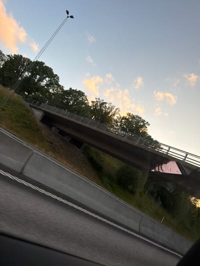 The image depicts an overpass on a highway in Munkedal, Sweden, during sunset. The sky is a pale blue with scattered clouds tinged with orange and pink from the setting sun. A tall, lattice-like lamppost with two lights stands prominently against the sky. Lush green trees line the embankment beneath the overpass, and the overpass itself has a metal railing. The highway, in the foreground, has a concrete barrier and double white lines painted on the asphalt, indicating a road in motion. The overall scene suggests a transitional moment of the day, with natural elements and infrastructure coexisting.