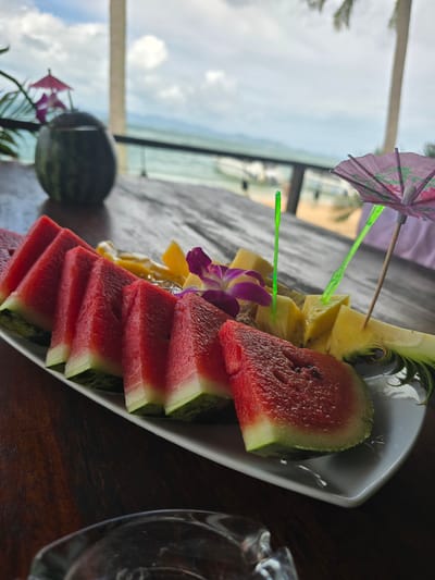 A close-up, skewed-perspective shot features a white platter filled with sliced watermelon, pineapple, and a purple orchid. The watermelon is stacked in slices along the platter's edge. Two cocktail umbrellas are inserted into the fruit. In the blurred background, a window reveals an ocean view under a cloudy sky, with a palm tree visible on the left. A partially visible glass object sits on a dark wooden surface in the lower right corner. The scene is suggestive of a tropical resort setting. The location is Ban Khlong Hia, Thailand.