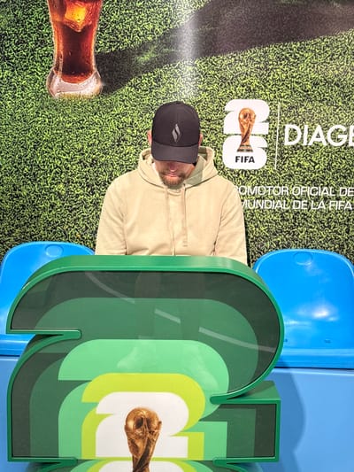 The image depicts a person wearing a black baseball cap and a light-colored hoodie, seated in front of a backdrop featuring a green, textured surface resembling grass, a glass of dark liquid with ice, a FIFA World Cup trophy, and text that includes "DIAGE", "FIFA", and "OMOTOR OFICIAL DE MUNDIAL DE LA FIFA". Two blue plastic seats are visible on either side of the person. In the foreground, a large, multi-layered green decorative object with a cutout resembling the number "2" is present, also featuring a smaller replica of the FIFA World Cup trophy. The scene appears to be indoors, possibly at an event or exhibition related to the FIFA World Cup, with the FIFA logo and text suggesting an official sponsorship. The specific location in Ezeiza, Argentina, is not discernible from the visual information provided in the image.
