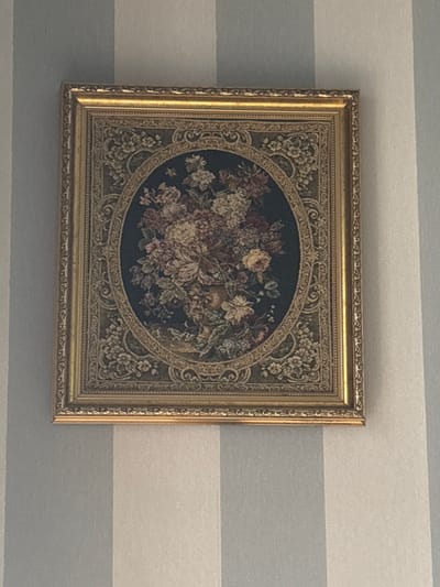 The image shows a framed tapestry with an oval floral design, hanging on a striped wall. The tapestry is richly detailed, featuring an assortment of flowers in various shades of pink, white, and yellow against a dark background. The oval center is surrounded by an ornate border with intricate scrollwork and floral motifs. The frame is gold-colored with decorative trim. The wall behind the tapestry has horizontal stripes in alternating light gray and white. The lighting suggests it is daytime, possibly indoors, and the overall mood is calm and decorative. There is no visible text in the image. The location is identified as Mińsk Mazowiecki, Poland.