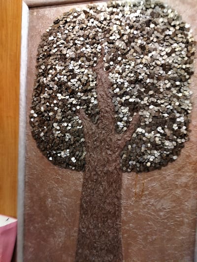 The image depicts a framed artwork, likely a relief or mosaic, that resembles a tree. The tree's trunk and branches are textured, suggesting a rough bark, and are made of a darker material, possibly clay or wood. The foliage of the tree is entirely composed of numerous small, circular objects that appear to be coins. These coins, varying in color from silver to bronze, are densely packed, forming a full, rounded canopy. The background of the artwork is a textured surface in shades of brown and beige, with some areas appearing smoother and others having a rough, almost rippled appearance. The artwork is framed with a light-colored, possibly wooden, border. To the left of the artwork, a portion of a wooden surface and a hint of a pink object are visible, suggesting a domestic or indoor setting. The lighting appears to be artificial, casting subtle shadows and highlighting the metallic sheen of the coins. No people are visible in the image. The artwork itself is the sole subject. The scene is static, with no activity taking place beyond the static display of the artwork. There is no visible text in the image. The provided location context of Staryi Aidar, Ukraine, does not offer any direct visual cues within the image itself.