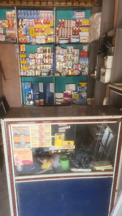 This image depicts the interior of a small pharmacy or drug store in Jogana, Nigeria. The store features shelves stocked with various medications and pharmaceutical products, arranged in rows and columns. Brands such as "Kufcof", "Lablin", "DOGOFEN", "SANAVITA", "On Call Plus", and "Neoskin" are visible. A glass display case contains items such as "KAKARAKA POWER" and "EXTRAJOSS". The presence of "SANAVITA RE-USE PREVENTION SYRINGES" and "RINGES 1x100" suggests a focus on medical supplies. Promotional materials and business cards are also visible inside the display case. The setting appears modest and typical of small retail establishments in developing urban areas.