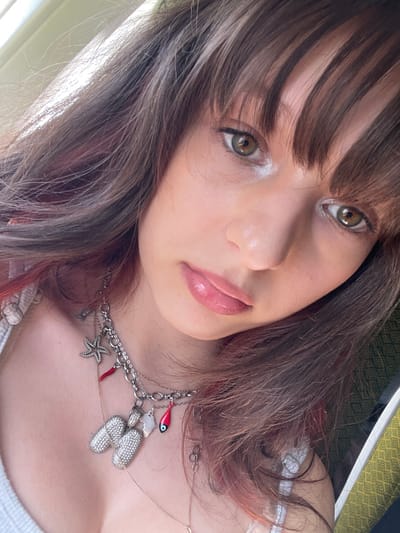 A close-up selfie captures a young female with long, brown hair featuring subtle reddish-pink highlights and blunt bangs. Her hazel/green-brown eyes are open and directed towards the camera, while her lips are full and glossy pink. She wears a light-colored, possibly white, top with a visible strap on her left shoulder. Adorning her neck are multiple necklaces: a prominent silver-toned chain adorned with various charms, including a large pave-set initial "M", a textured starfish, a red chili pepper (cornicello), and a red fish charm detailed with an evil eye motif. A thinner, delicate gold-toned chain is also visible beneath the main necklace. The background is softly blurred, revealing a bright light source on the upper left and a hint of a greenish-yellow patterned textile or surface on the right. The scene is located in the city of Tzafria, Israel.