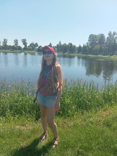 A full-shot, outdoor, medium-light skin toned woman wearing a red baseball cap, sunglasses, a multi-colored sleeveless top, and a light blue denim skirt stands in a grassy area with tall green grass in the foreground. She is facing slightly left with her head tilted back and her mouth open in a smile. Behind her is a large body of water, likely a lake or river, with trees and a fence line along the far bank under a clear blue sky. The scene is set in the city of Wasilków, Poland.