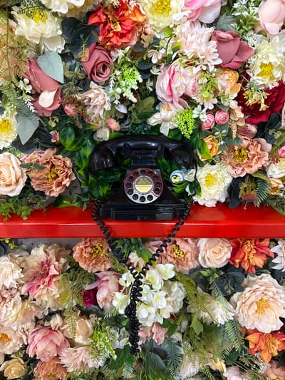 The image presents a vibrant, static display featuring a vintage black rotary telephone set against a dense backdrop of artificial flowers and greenery. The scene is dominated by an abundance of faux blooms in a rich palette of colors, including various shades of pink, cream, white, deep red, orange, peach, and yellow, intermingled with lush green leaves and fern-like foliage.
A classic black rotary telephone is centrally positioned, with its handset detached from the cradle and hanging by its coiled cord. The phone's body and dial are nestled amongst the flowers on the left side of a prominent, glossy, bright red vertical strip that runs down the middle-right of the composition. The rotary dial features a central yellow disc where small black text is visible, indicating emergency numbers: "FIRE - 0", "POLICE - 02", and "AMBULANCE - 03".
The floral arrangement is meticulously crafted, creating a full and textured wall of blossoms that surrounds the telephone and extends beyond the frame. The lighting is bright and uniform, enhancing the vivid colors and details of the artificial plants and the vintage phone. The setting appears to be an art installation or decorative wall, possibly indoors in Korobovo, Russia, evoking a sense of whimsical charm and nostalgia. No human subjects or specific activity are present.