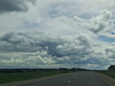The image shows a paved road stretching into the distance through a rural landscape. On the left side of the road is a green field. On the right side of the road, the terrain slopes upwards and is covered with grass. In the distance, there are trees forming a dense forest line along the horizon. Above the landscape, the sky is filled with dramatic, heavy clouds in varying shades of gray and white, suggesting an overcast or possibly stormy weather condition. A few low-lying buildings and structures, possibly an industrial facility or farm complex, are visible in the mid-ground on the left side, nestled amongst the trees. The road itself has white dashed lines indicating lanes. The overall impression is a serene, yet somewhat imposing, natural scene with man-made elements present but not dominating the landscape.