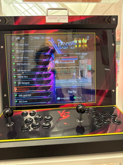 The image shows an arcade cabinet with a digital display and a control panel. The display shows a game menu for "Pandora Saga DX Plus" with a list of game titles and a game in progress. The control panel features two joysticks and several buttons. A sign at the top of the cabinet reads "$50 CENTS TO PLAY". The arcade cabinet is positioned in front of a retail display with fluorescent lighting overhead. The scene appears to be indoors, likely in a store or arcade, and is located in El Paso, United States of America. There are no visible people in the immediate vicinity of the arcade cabinet. The overall impression is that of a typical arcade setup.