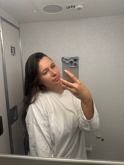 A person is captured in a mirror selfie, holding a smartphone to take their own picture. The image is oriented vertically, as if the camera was rotated 90 degrees clockwise from a standard landscape shot.
The subject is a woman with long, dark brown hair, which is parted on her left side and cascades over her left shoulder and down her back. She has a fair complexion with some minor blemishes visible, and her eyebrows are dark. Her eyes are looking directly towards the phone, and her lips are slightly parted, revealing a neutral or contemplative expression. She appears to be wearing a plain, light-colored (likely white or off-white) long-sleeved t-shirt or top, which is loose-fitting and shows some wrinkles.
Her right hand holds a gray smartphone, which appears to be a modern iPhone model with a prominent triple-lens camera module. The phone is encased in a distinctive, light gray, irregularly shaped case that features a wavy or organic design element on the left side and a clear cut-out for the cameras. Her fingernails are painted a light, subtle color, possibly pink or nude. On her right ring finger, she wears a stack of at least two thin, metallic rings.
The setting appears to be an interior space with light gray, speckled or textured walls, possibly resembling a small room like an airplane or train lavatory, or a compact cabin space. In the upper left portion of the reflection, a white vent or air conditioning unit is visible, with a round speaker-like grill directly beneath it. The bottom left of the reflection shows what looks like a metallic handle or latch, possibly part of a door. The right edge of the mirror frame is visible, appearing white or light-colored. The lighting is even, suggesting indoor artificial light. No discernible text is visible anywhere in the image.