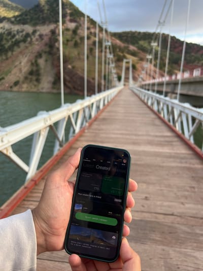 The image shows a person standing on a bridge, holding a smartphone in their hands. The phone displays a mobile application related to finances and content creation. The person is wearing a white long-sleeved shirt. The bridge has a wooden deck with steel supports and is set against a backdrop of mountainous terrain and water.
