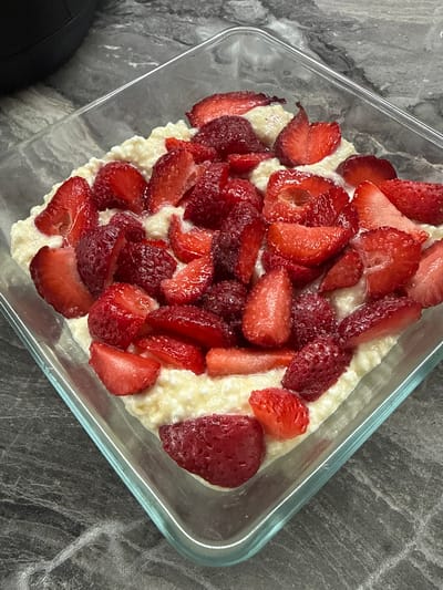 A square glass bowl filled with cottage cheese topped with sliced strawberries sits on a dark gray marble countertop. A black object is partially visible in the upper left corner of the frame. The cottage cheese is a creamy white color, and the strawberries are bright red and glistening. The image is taken from a slightly elevated angle, looking down into the bowl. The lighting is natural, casting soft shadows. This scene is likely a home kitchen in Chakvi, Georgia.