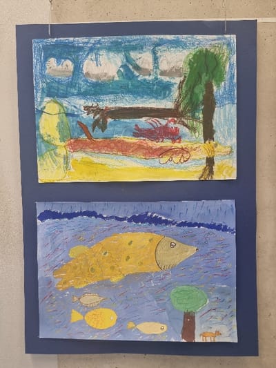 The image displays two horizontally oriented, unframed children's drawings, created with crayons or pastels, mounted vertically on a dark blue backing sheet. This assembly is suspended by thin wires against a light grey, textured wall in Jaunmārupe, Latvia.
The upper drawing depicts a simplified landscape. The top third features a light blue sky with several irregular grey and white cloud formations. Below the sky is a strip of dark blue, representing water. The foreground comprises a vivid yellow ground, likely sand or land. A dark green tree with a brown trunk is positioned on the right side, extending from the yellow ground into the blue water area. On the yellow ground, left of the tree, a red, multi-legged creature resembling a lobster or crab is visible. Above this creature, a long, dark brown horizontal shape extends. An amorphous light green shape occupies the far left of the yellow ground.
The lower drawing illustrates an aquatic scene with a small terrestrial element. The upper section of the drawing features a dark blue area with white wavy lines, suggesting the water's surface or deeper water. The primary water body is a lighter blue, filled with numerous short, multicolored crayon strokes in red, yellow, green, and blue, representing water currents or movement. A large, golden-yellow fish with a light brown head and distinct eye is centrally located, facing right, with small circular textural details on its body. Bubbles are depicted emanating from its mouth. Below this large fish, three smaller fish of varying sizes and beige-yellow coloration swim towards the left. In the bottom right corner, a green, leafy tree with a brown trunk is depicted on a small patch of brown ground. Adjacent to the tree's base, a small, four-legged brown animal, consistent with a fox or dog, stands.