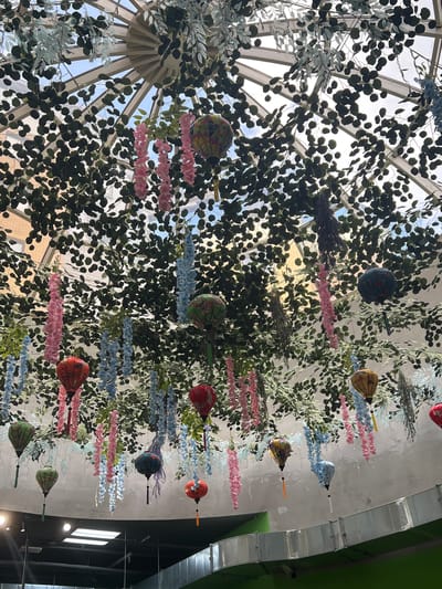 The image depicts an interior view of a structure with a glass dome ceiling, possibly a conservatory or atrium. Through the glass, hints of the sky can be seen. Dense foliage and decorative elements, including lanterns and hanging floral garlands, are suspended from the ceiling. The decorations are predominantly pink and blue. Ductwork runs along the interior architecture. The specific location is Вёшки, Russia.