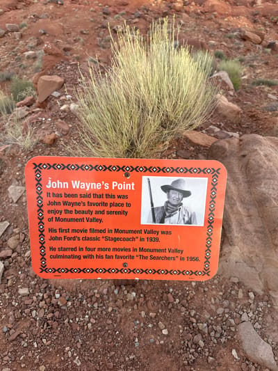 An orange sign with black and white geometric borders stands in a rocky, desert environment. The sign reads "John Wayne's Point" and states that it was his favorite place to enjoy the beauty and serenity of Monument Valley. It also mentions his first movie filmed in Monument Valley was "Stagecoach" in 1939, and that he starred in four more movies there, culminating in "The Searchers" in 1956. A black and white photograph of John Wayne in cowboy attire holding a rifle is displayed on the right side of the sign. The background consists of red rocks, sparse vegetation, and gravel. The location is Oljato-Monument Valley, United States.