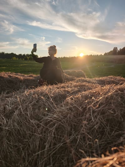 A wide outdoor shot captures a young woman sitting on a large hay bale in a sun-drenched field in Стеклянный, Russia, at what appears to be sunset or golden hour. The woman, positioned towards the left side of the frame, is holding a smartphone in her raised left hand, pointing it towards the distant landscape, suggesting she is taking a photo or a selfie. She is seen in profile, wearing dark clothing, possibly a hooded top, with her hair tied up. The warm, golden light of the setting sun illuminates her from behind, creating a partial silhouette and a soft glow around her form.

The sun itself is a bright, intense orb on the right side of the image, resting just above a distant treeline, and casts prominent lens flares into the scene. The sky is a blend of pale blue and soft orange, adorned with delicate, wispy white clouds that stretch across the upper portion of the frame. Below the treeline, a green field extends, indicating a rural or pastoral setting. The foreground is dominated by the intricate textures of dried hay bales, with the straw catching the warm light, adding depth and a rustic feel to the composition. The overall mood is peaceful and scenic, emphasizing the beauty of the natural landscape bathed in the late afternoon sun. No visible text is present in the image.