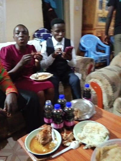 Two individuals are seated and enjoying a meal. The person on the left wears a red dress and is eating. The other person, dressed in a white shirt and dark vest, is focused on a mobile phone. A low table in front of them is laden with several bottles of soda, plates of food, and used cutlery. A blue plastic chair is slightly visible in the background. The setting appears to be indoors.