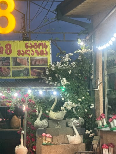 The image depicts an outdoor commercial establishment in Manavi, Georgia, at dusk or night, illuminated by artificial lights. A dark blue sky is visible in the upper portion, featuring a sparse metal framework with wires and some climbing green foliage. A large, bright yellow, illuminated number "8" is prominently displayed on the upper left. Below it, a yellow banner with red Georgian text reads "ნატურალური პროდუქტები" (Natural Products), flanked by another red "8" on the left. The banner features six grid-like sections displaying various food products, including what appear to be sausages or churchkhela, and possibly dried fruits or vegetables.
The foreground and midground are densely decorated with white and red climbing roses. Multiple strings of warm white, round-bulb lights are strung across overhead structures and along the facade. Additional spotlights, including one emitting a green light on the left, illuminate the foliage and sculptures.
Several decorative elements are present:
* Four light-colored swan sculptures are arranged at various levels on concrete or stone pedestals and steps in the central and right areas of the image. The swans vary slightly in size and pose.
* Multiple red and white mushroom sculptures are visible, with some arranged on a shelf against a building facade on the right, and others positioned on the lower steps.
* On the far left, partially obscured by red roses, a large, dark, tear-drop shaped object, possibly a decorative pot or lamp, rests on a surface.
The building facade on the right consists of textured brown and tan sections with a glass window reflecting some lights. A small security camera is mounted on the wall amidst the white roses. No people are visible in the scene. The overall impression is that of a richly decorated, inviting outdoor space, likely a shop or restaurant, emphasizing natural products.