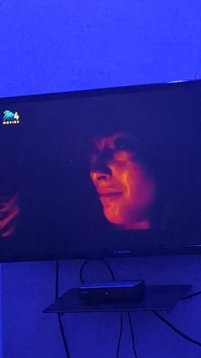 A television displays a dark image of a man's face in red and orange hues. The background appears a deep blue. A smaller black rectangular device is attached to the back of the TV, wires extending behind it. In the lower left corner of the screen, there is a logo for a movie channel. The overall scene is dimly lit, emphasizing the colors on the screen.