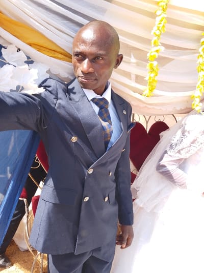The image depicts an outdoor celebratory scene in Angwan Sarki, Nigeria. A bald, dark-skinned adult male is positioned centrally, facing slightly left of the camera's axis with a serious expression. He wears a dark blue double-breasted suit featuring six gold buttons, a white collared shirt, and a blue and gold patterned necktie. A blue pocket square with gold trim is visible in his left breast pocket, along with a small gold lapel pin on his left lapel. His left arm is extended forward, suggesting he is holding the camera. To the male's right, a partial view of an individual in a white garment with lace detailing, consistent with a wedding dress and veil, is visible. The background features draped fabrics in white, gold, and blue, some with patterns. Yellow floral garlands hang vertically on the right side of the frame. A large, dark red heart-shaped decoration is partially visible behind the male subject. The ground in the lower-left foreground appears to be light brown soil. The scene is illuminated by bright natural light.
