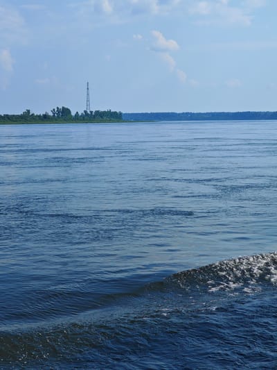 The image captures a wide, calm river on a partly cloudy day. The water is a deep blue, reflecting the sky. In the distance, a tree-lined shore stretches across the horizon, dotted with a tall, lattice-like communication tower on the left side of the frame. The foreground shows a small wave or wake breaking, creating white foam against the darker blue water. The scene appears to be a natural landscape with some man-made infrastructure in the distance.