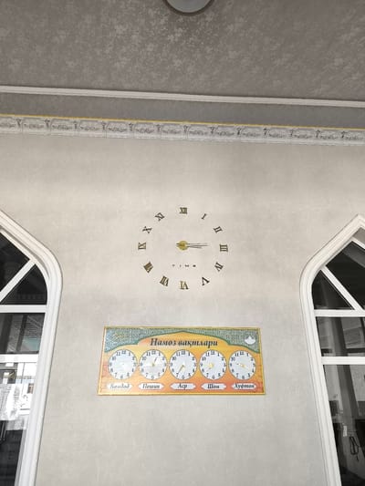 The image shows the interior of a building with a textured light gray wall and ceiling. A decorative white molding with gold accents runs along the top of the wall. To the left and right are arched windows with white frames. Centered on the wall is a large Roman numeral clock with a gold clock hand. Below the clock is a rectangular plaque displaying five smaller clocks, each with a different prayer time indicated. The plaque has the title "Намоз вактлари" (Prayer Times) in Cyrillic script at the top. The prayer times are labeled in Cyrillic below each clock: "Бомдод" (Bomdod), "Пешин" (Peshin), "Аср" (Asr), "Шом" (Shom), and "Хуфтон" (Hufton). The environment suggests this is a place of worship or a community center in Бабаходжа, Uzbekistan, where prayer times are prominently displayed. There are no people visible in the image.
