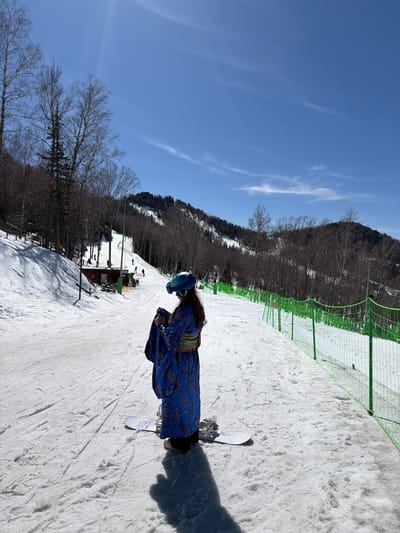 The image depicts a person snowboarding on a snowy slope. The person is wearing a blue patterned garment and a helmet. The surrounding environment includes a forest, a ski resort fence, and a clear blue sky. There is no indication that the location is in the city of Ozernoe, Russia.