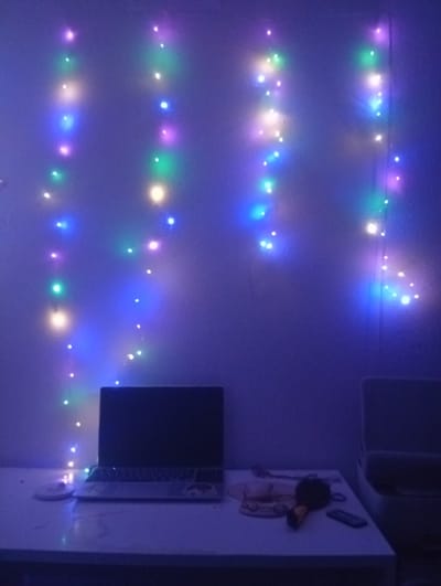 The image depicts a dimly lit room, likely a bedroom or study, with a white desk in the foreground. On the desk, a silver laptop is open, its screen dark. To the right of the laptop are several small objects, including what appears to be a remote control, and a small, fuzzy black item. The wall behind the desk is decorated with strings of colorful fairy lights, casting a soft glow in shades of blue, green, purple, and yellow. The lights are arranged in vertical strands, creating a festive atmosphere. The overall scene is calm and tranquil. The image does not contain any information or visual cues that would allow for the determination of its location in the city of Viviers, France.