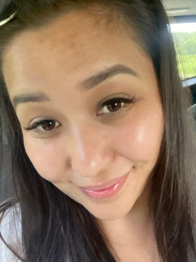 This is a close-up selfie of a woman with long, dark hair, taken outdoors during the daytime. She is smiling and looking directly at the camera. Her eyes are brown, and she is wearing mascara and eyeliner. Her lips are glossy with pink lipstick. The background is blurred, but appears to be a natural setting with green foliage, suggesting she is in a park or wooded area. There is no indication of the specific location within the city of Tyulyugul, Russia.