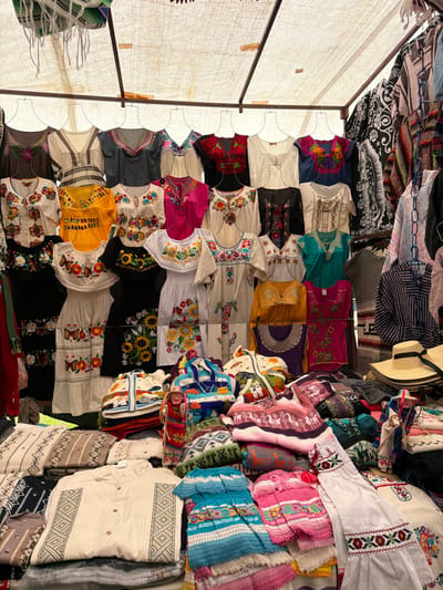 This image captures a vibrant and densely packed market stall, overflowing with a wide array of colorful textiles and traditional clothing. The scene is viewed from slightly below, looking up into what appears to be a covered booth, possibly under a tent or awning.
Overhead, a light-colored, possibly off-white or beige, fabric canopy stretches across the top and left side of the frame, providing shade. Support poles for this canopy are visible on the left.
The primary focus of the image is the extensive collection of garments, mostly blouses and dresses, arranged on hangers and stacked in piles.
On the left and center, numerous blouses are displayed on hangers. These garments feature rich, intricate embroidery in various styles. Many are white, black, or colored (pink, blue, yellow, green) with vibrant floral patterns, including sunflowers, roses, and other blossoms, executed in bright reds, yellows, blues, and greens. Other blouses show geometric embroidery or abstract designs around the necklines and sleeves. The styles suggest traditional Mexican or Latin American folk attire, such as huipiles.
Towards the upper right, several items are hanging or draped, including blankets or shawls with striped patterns and dark fabrics featuring white, intricate designs. A light-colored straw hat with a dark band is also visible, hanging in this section.
On the right side of the stall, large piles of folded textiles are neatly stacked. These appear to be thicker woven garments, possibly sweaters, ponchos, or blankets, showcasing a diverse range of patterns and textures. Colors include various shades of pink, purple, blue, green, and cream, often with complex geometric motifs or stylized animal patterns woven into the fabric. Additionally, there are several cream-colored tunics or shirts with distinct geometric embroidery along the front and hems.
The bright, natural lighting suggests it is daytime. No people are visible in the frame, and no discernible text appears on any of the items or in the background. The overall impression is one of a bustling and colorful artisan market, rich in cultural textile traditions.