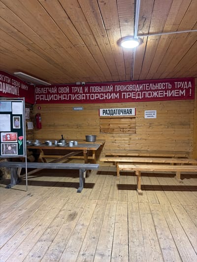 The image shows the interior of a rustic, wooden room, possibly a canteen or communal dining area in Ust'-Pyzas, Russia. A large red banner with white Cyrillic text stretches across the upper portion of the wall, reading "Облегчай свой труд и повышай производительность труда рационализаторским предложением!" (Make your work easier and increase labor productivity with a rationalization proposal!). To the left, another banner with red text on a lighter background says "КУПИ СВОЮ ВИНУ ОДИННОЙ!" (Buy your wine, single!).
In the center of the room, a long wooden table is set with numerous metal pots, pans, and cups, suggesting it's a serving area. A large metal kettle sits prominently on the table. To the right of the table, two wooden benches are positioned against the wall. A sign above the benches reads "РАЗДАТОЧНАЯ" (Serving area). Another small sign to the right of this one states "Экспонаты руками не трогать" (Do not touch exhibits with your hands).
On the left side of the image, a display board is set up with framed photographs and documents. A red fire extinguisher is mounted on the wall behind the table, near the display board. The room is illuminated by a ceiling light fixture. The walls and ceiling are constructed from wooden planks, contributing to the rustic aesthetic. The floor is also made of wooden planks. The overall impression is of a simple, functional space, likely part of a historical or industrial site. There are no people visible in the image.