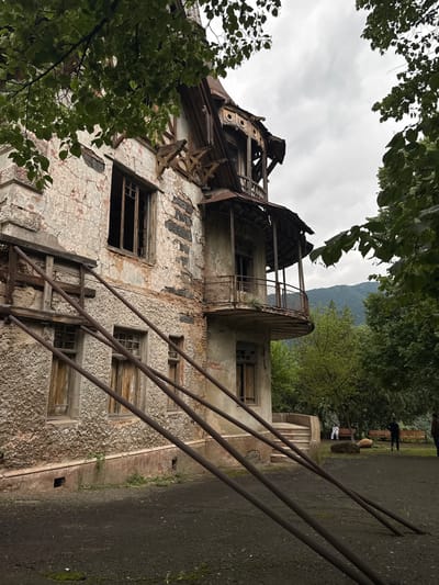 An abandoned building with a decaying facade stands amidst lush greenery, with a cloudy sky overhead. The building exhibits signs of severe disrepair, with peeling plaster, broken windows, and sections of exposed brick. A rusted metal railing system is precariously attached to the exterior. In the background, several trees are visible, and further in the distance, a mountain range can be seen under the overcast sky. To the right of the building, a set of concrete stairs leads up to an entrance. Several people are present in the background, some seated on benches and others standing. The ground in the foreground is covered in dark gravel.