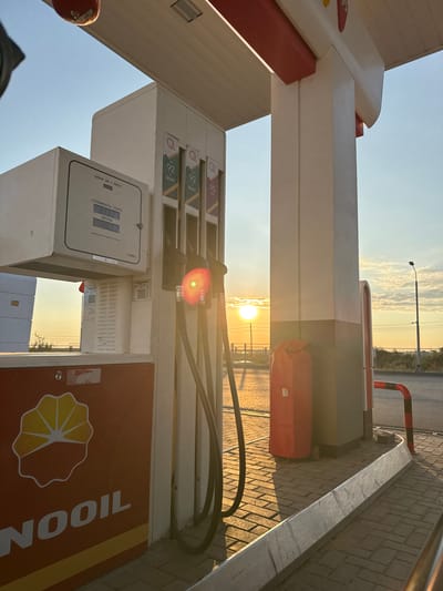 The image depicts a gas station at sunset in the city of Belkol, Kazakhstan. The focal point is a white and beige gas pump with three nozzles visible. The pump has digital displays showing prices and volume in liters. The "NOOIL" brand logo is prominently displayed on the lower part of the pump. In the background, the sun is setting, casting a warm glow and long shadows. The sky is partly cloudy and a street lamp is visible in the distance. The ground is paved with interlocking bricks, and a red trash can on wheels is positioned next to the pump.