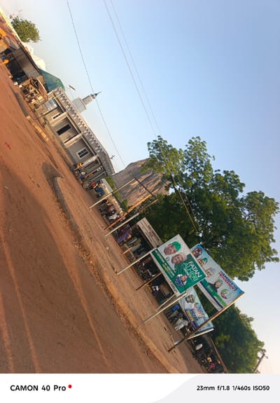 The image shows a street scene in Wase, Nigeria. The foreground is a dirt road running diagonally across the frame. There are several political billboards along the right side of the road, featuring images of candidates. In the middle ground, there is a simple building with a minaret, suggesting it is a mosque. People are gathered near the billboards. In the background is a rocky mountain or hill. The sky is clear and blue. Power lines are visible in the upper part of the image.
