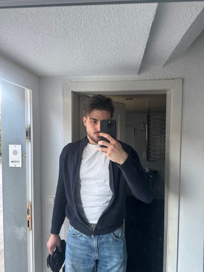 The image displays an adult male taking a mirror selfie in a hallway or entryway, identified as being in the city of Rafz, Switzerland. The male has dark hair and stubble, and is wearing a white t-shirt, an unzipped dark blue ribbed knit cardigan, and light blue denim jeans. He holds a dark-colored smartphone in his left hand, covering a portion of his face. His right hand holds an indistinct dark object. The environment reflected in the mirror includes a white textured ceiling with a recessed panel. To the left side of the reflection, a doorway leads outdoors or to a brightly lit area, showcasing an external view with sunlight. A white rectangular intercom system and gold-colored door hardware are mounted on the wall adjacent to this doorway. To the right side of the reflection, another doorway reveals an interior room containing a chrome-colored horizontal-slatted towel warmer or rack mounted on a wall. The surrounding walls are light grey.