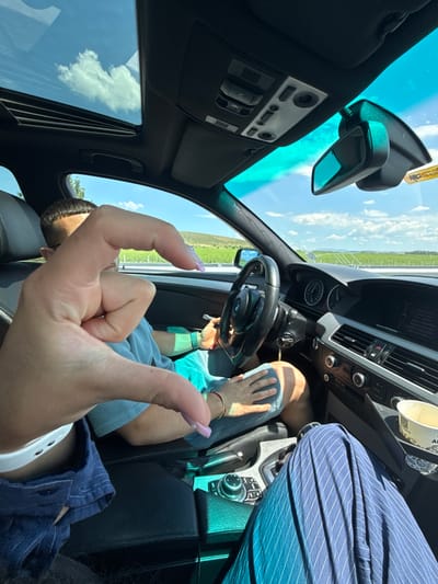 In Kermen, Bulgaria, two individuals are seated inside a car with its sunroof open, revealing a bright, cloudy sky. The driver, a man with short dark hair, is in the driver's seat, holding the steering wheel. In the foreground, a hand with light pink nail polish forms a heart shape. The interior of the car shows a dashboard with a navigation screen, air vents, and various controls. Outside the car, a green landscape with rows of plants, possibly vineyards, stretches towards rolling hills under a blue sky. The scene appears to be a scenic drive on a sunny day.