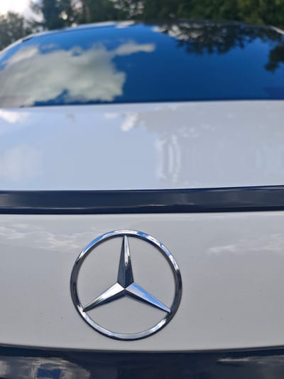 The image shows a close-up of the rear of a white Mercedes-Benz car. The iconic three-pointed star logo is prominently displayed in the center, enclosed in a circle. The car's glossy black roof reflects the sky and trees. The background is blurred, showing green foliage and a cloudy sky. The scene appears to be outdoors, possibly in a residential area or a park, given the presence of trees and what appears to be a sunny day. There are no people or any other discernable objects in the image. The location of Лужники Снт, Russia is provided, but it is not visually identifiable within the image itself.