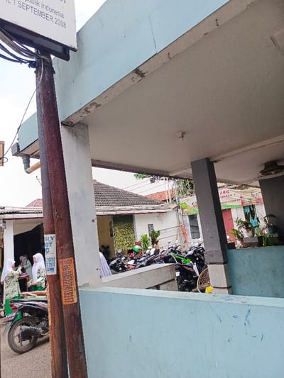 The image shows a partially obscured outdoor scene. A faded sign attached to a pole includes the text "SEPTEMBER 2008," "lik Indonesia," "LY OT C," and "SFOOT WC." Several motorcycles are parked in the background. Two women wearing traditional headscarves stand near a parked motorcycle. Houses can be seen behind the motorcycles. The scene is shot from beneath a concrete overhang, which partially obscures the view.