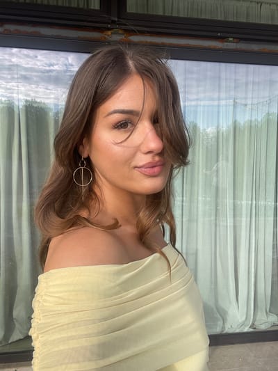 A woman is photographed indoors in a vertical composition. The woman has light skin, brown hair with highlights, and a light yellow strapless dress. She is wearing a gold earring with a circular design. The background consists of a window with a view of a sky and trees, obscured by a light-colored curtain. The window frame is dark-colored. The location is unspecified and there is no indication this is in Беране, Montenegro.