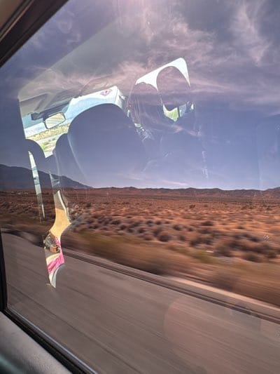 The photograph shows a view from inside a car moving through a desert landscape. The interior of the car is reflected in the window, showing the back of the driver's seat, the headrest, and the rearview mirror. A person is partially visible in the reflection as well. The landscape outside the window consists of a straight road running through a desert environment with sparse vegetation and distant mountains. The sky is visible above, with a mix of blue and light clouds. The blurring suggests the car is in motion. The city of Desert Hot Springs, United States is not directly identifiable from the image.