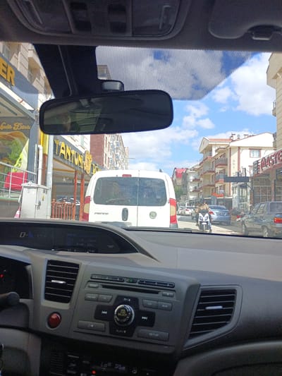 The image is taken from inside a vehicle, looking forward through the windshield. The scene is a street in Yahşihan, Turkey, on a cloudy day. In the foreground, the dashboard of the car is visible, including the steering wheel, air vents, and the car's entertainment system. Outside the car, traffic is visible. A white Fiat Fiorino van is directly in front, and further down the street, a person is riding a scooter. The buildings lining the street appear to be residential or commercial. A restaurant with a yellow awning, identified by a sign reading "TANTUNİ," is on the left side of the street.