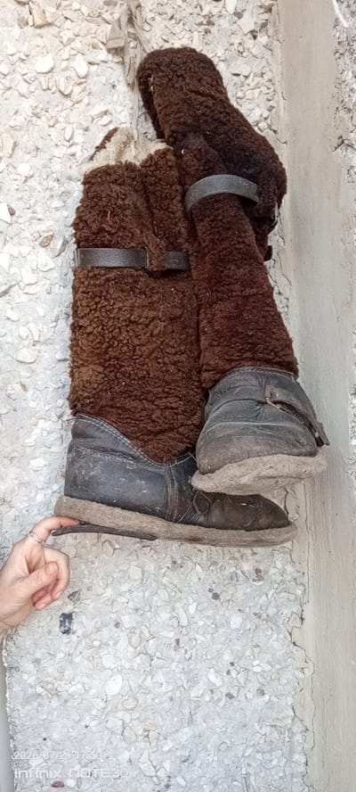 A pair of dirty, brown, fur-lined boots are hung on a rough, speckled concrete wall. The boots appear to be made of leather and have straps across the top. A person's hand with a ring on their finger is touching the sole of the lower boot. The background is a textured wall made of concrete with small stones embedded in it. The lighting suggests it is daytime. The image appears to have a date stamp in the bottom left corner indicating July 29, 2026, and "Infinix NOTE 30." No specific location in Langepas, Russia, can be identified from the image.