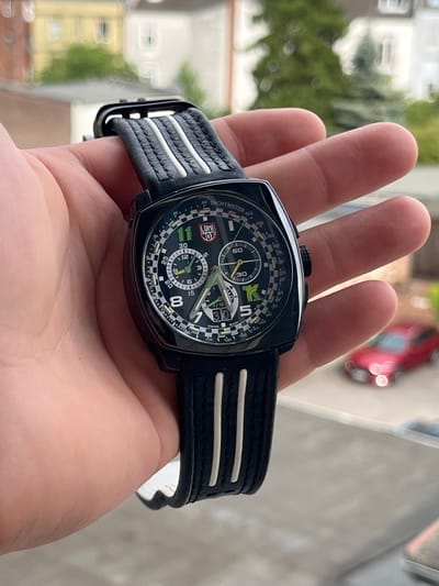 A close-up shot shows a person's hand holding a black Luminox watch with a black strap. The watch face is round with a black background and green and white accents. The watch features multiple subdials and a tachymeter scale. The strap has three white stripes. In the blurred background, a red car is visible, suggesting an outdoor setting possibly in Neuss, Germany. The lighting indicates it is daytime, with a soft, diffused light. The focus is on the watch, highlighting its design and details. The image appears to be a product shot or a personal photo showcasing the timepiece.