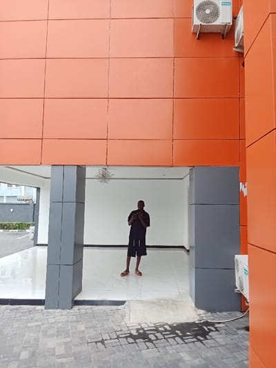 The image shows a man standing in a doorway. He is wearing a black long-sleeved shirt, black shorts, and brown sandals. His hands are in front of him, holding something. The doorway is flanked by two large grey pillars, and the wall above is orange. The floor is tiled in a white and grey pattern. An air conditioner is mounted on the orange wall. This scene appears to be in a modern building. The location is the town of Ujevwu, Nigeria. It is at latitude 5.472, longitude 5.817.
