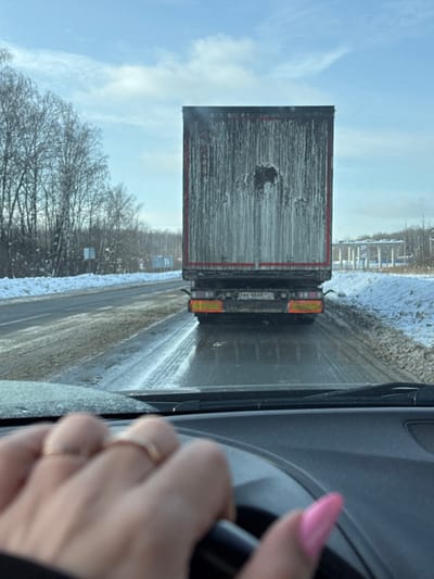 The image presents a driver's perspective from inside a vehicle on a winter road in L'vovskiy, Russia. In the bottom-left foreground, a light-skinned hand, exhibiting two rings on the ring finger and pink-painted fingernails, grips a black steering wheel. Directly ahead, a dark semi-trailer occupies the central frame, viewed from the rear. The trailer's surface is heavily streaked with a pale, possibly frozen or dried, residue. A Russian license plate, "РВК 4648 77," is affixed to the lower rear of the trailer. The asphalt road surface appears wet and reflective, with visible tire tracks, indicating moisture or icy conditions. Snow covers the ground along the roadside verges. To the left, a line of bare deciduous trees and some evergreens are visible, partially covered in snow. The sky is predominantly clear blue with sparse white clouds. In the distant background on the right, an elevated white structure, consistent with a gas station canopy or a toll booth, is discernible.