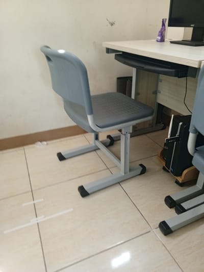 The image depicts an indoor scene, likely a classroom or office, with a focus on a grey plastic and metal chair and a desk. The chair is positioned in the foreground, to the left of the frame, with its back facing the camera at an angle. The desk is visible in the upper right portion of the image, supporting a computer monitor and keyboard. A small purple bottle is on the desk. The floor is tiled with beige square tiles. The walls appear to be painted a light color. There are no people visible in the image. The scene is static and does not suggest any particular action or interaction. The environment is sparsely furnished and functional. The specific location within Chutula, Mozambique, cannot be determined from the visual information provided.