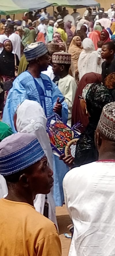 The image captures a densely crowded outdoor gathering in Sani Mai Nagge, Nigeria. Numerous individuals, predominantly adults, stand closely together on unpaved ground under bright daylight conditions. Many men wear traditional patterned caps and long robes (boubous), while women are largely observed in head coverings, including hijabs and veils, alongside varying traditional attire.
In the mid-ground, a man prominent in a light blue patterned boubou and a light blue patterned cap faces right, his right hand raised in a gesture of communication. To his immediate right, a younger person in a cream-colored garment and a patterned cap faces the gesturing man. Adjacent to this pair, another individual, partially obscured, holds a circular, multi-colored decorative object adorned with geometric patterns. In the immediate foreground, a person in an orange t-shirt and a blue-and-white striped cap is visible in profile, facing right. The background features a multitude of other individuals and several temporary structures, including fabric awnings or tents, one displaying green and white stripes, suggesting a market or public event setting.
