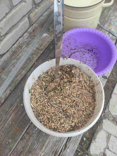 An overhead shot shows a white bucket filled with birdseed. A metal spoon with a floral pattern is sticking out of the seed. Behind the bucket is a purple plastic bowl with some dark residue inside. A yellow pot with handles is visible in the upper right corner. A metal ruler with inch markings lies diagonally on a wooden surface to the left of the bucket. The scene is set outdoors on a wooden table, with a brick wall in the background. There are no people present.