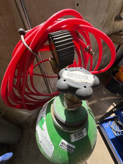 A green gas cylinder, identified by the label "MESSER SVAROVCI ARGON", is standing upright on a concrete surface. A thick red hose is coiled around the top of the cylinder, secured by a thin rope. A black regulator with a large, ribbed dial is attached to the valve of the cylinder, and a label with a barcode and the text "MUL 362214" is affixed to it. Another label on the cylinder reads "57787940". The environment appears to be an outdoor or semi-outdoor area, possibly a workshop or storage space, with a concrete floor and a textured wall in the background. The lighting suggests it is daytime, with sunlight casting shadows. There are no people visible in the image. The overall scene depicts equipment used for welding or gas supply.