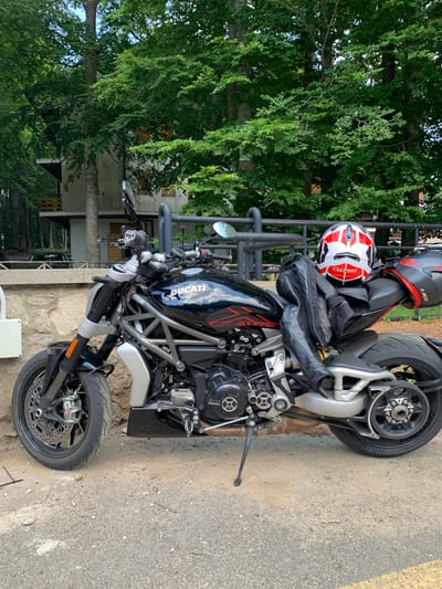 This media file features a black Ducati motorcycle parked on a gravel and asphalt surface. Two helmets, one red and white with a black stripe, and another predominantly black with red accents, are placed on the motorcycle's seat. A black leather jacket is draped over the seat as well. The motorcycle itself is detailed, with a visible "DUCATI" logo on the fuel tank and "BREMBO" branding on the brake caliper. The background includes lush green trees and a building with wooden elements, suggesting an outdoor setting. The image is taken during the daytime, with sunlight casting shadows. The scene appears calm, with no people visible, and the overall impression is that of a parked motorcycle ready for a ride. The location context is Seggiano, Italy.