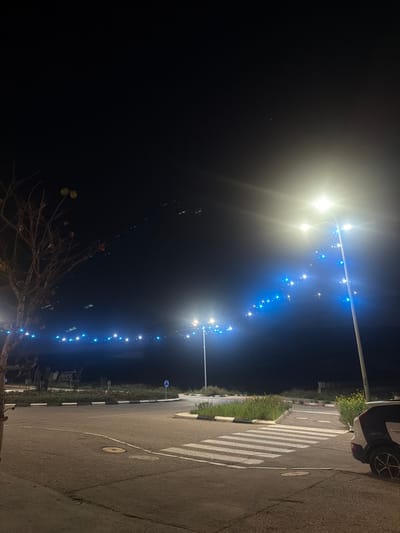 A nighttime view features a row of streetlights stretching into the distance, adorned with blue fairy lights. The right side reveals a paved road with a white-striped crosswalk, bordered by a strip of grass. A portion of a parked car is visible. A bare tree occupies the bottom left corner, contrasting with the illuminated background. The scene is captured in a slightly rotated perspective, creating a dynamic visual.