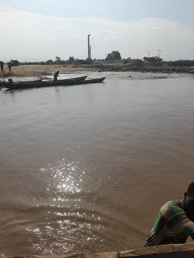 In Nemnei, Nigeria, a wide, muddy river occupies the foreground, reflecting the hazy sky and a bright sun. Several long, narrow wooden boats are moored along the sandy bank on the left side of the image. One person is visible in one of the boats. Further inland, construction equipment, including a large drilling rig, excavators, and piles of pipes, suggests ongoing development. Silhouetted figures of people are scattered across the sandy area. Power lines stretch across the background, with trees and buildings visible in the distance. In the lower right corner, a person wearing a horizontally striped shirt is partially visible, leaning on a wooden structure.
